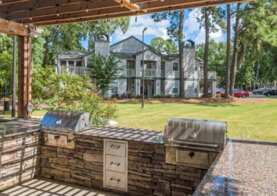 An outdoor kitchen with two grills and built-in cabinets, overlooking a grassy backyard with tall pine trees and a multi-story residential building.