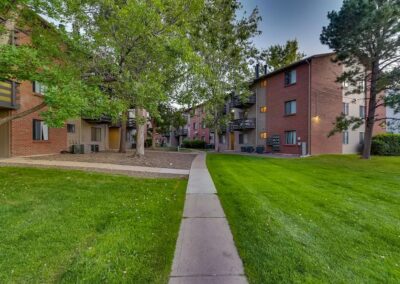 A well-manicured lawn leads to a pathway between multi-story brick apartment buildings with lush green trees in a serene residential area.