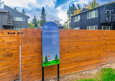 A wooden fence with an informational sign for a dog park named "aster park," surrounded by modern townhomes under a cloudy sky.