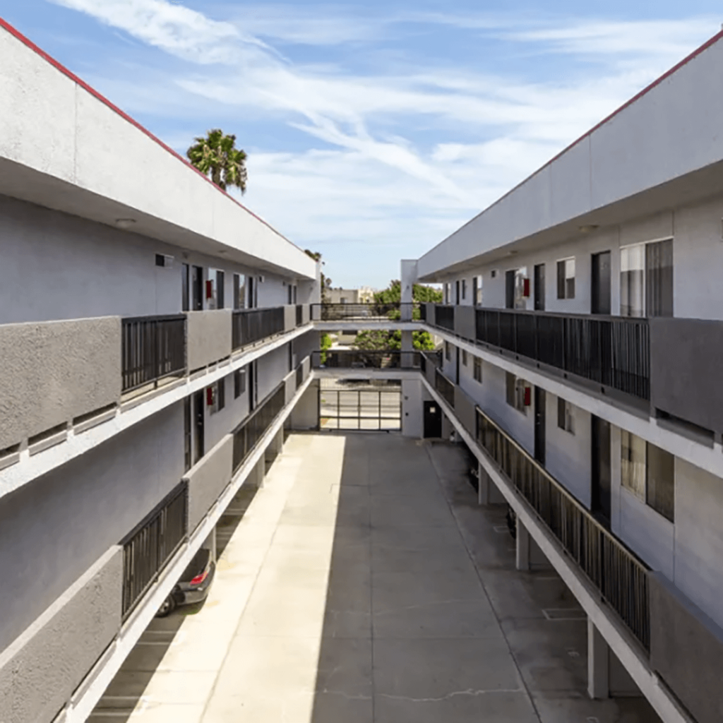 Symmetrical view between two apartment buildings with balconies, overlooking a central walkway under a clear blue sky, with a few palm trees visible in the background.