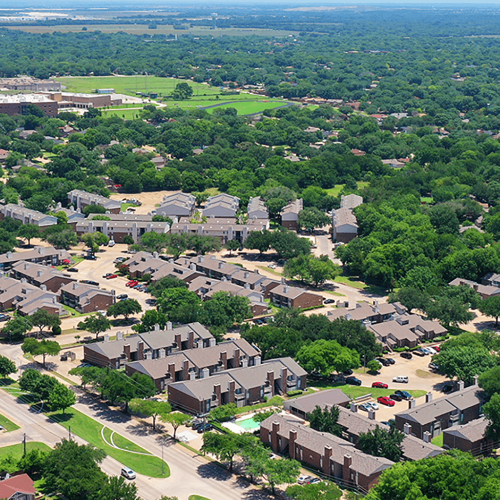 Aerial view of a suburban neighborhood with uniform brown-roofed houses, lush green trees, and winding roads, under a clear blue sky.