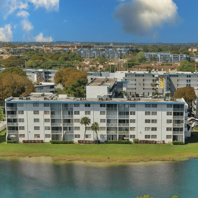 Aerial view of a modern, multi-story white apartment building beside a calm blue lake surrounded by greenery under a clear sky.
