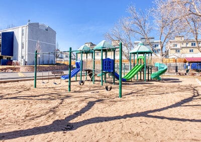 A sunny playground with multiple structures including slides, swings, and climbing frames, set in a sand area surrounded by trees and a fence, with buildings in the background.