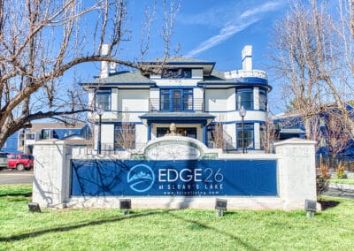 A two-story traditional house painted in blue and white, with a large promotional banner in front reading "edge 26 at sloan's lake" and a website url, located on a sunny day with clear blue skies.