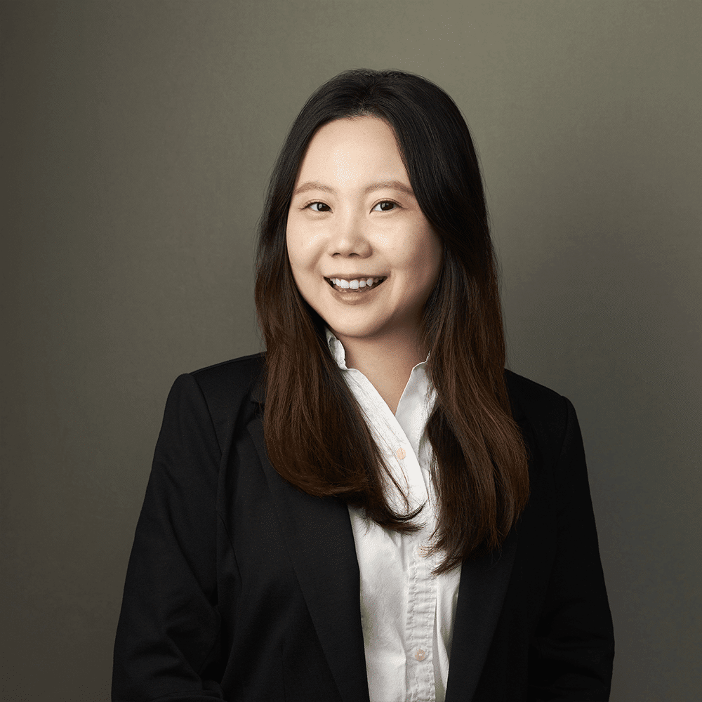 A professional portrait of a smiling asian woman in a black blazer and white shirt, set against a muted gray background.