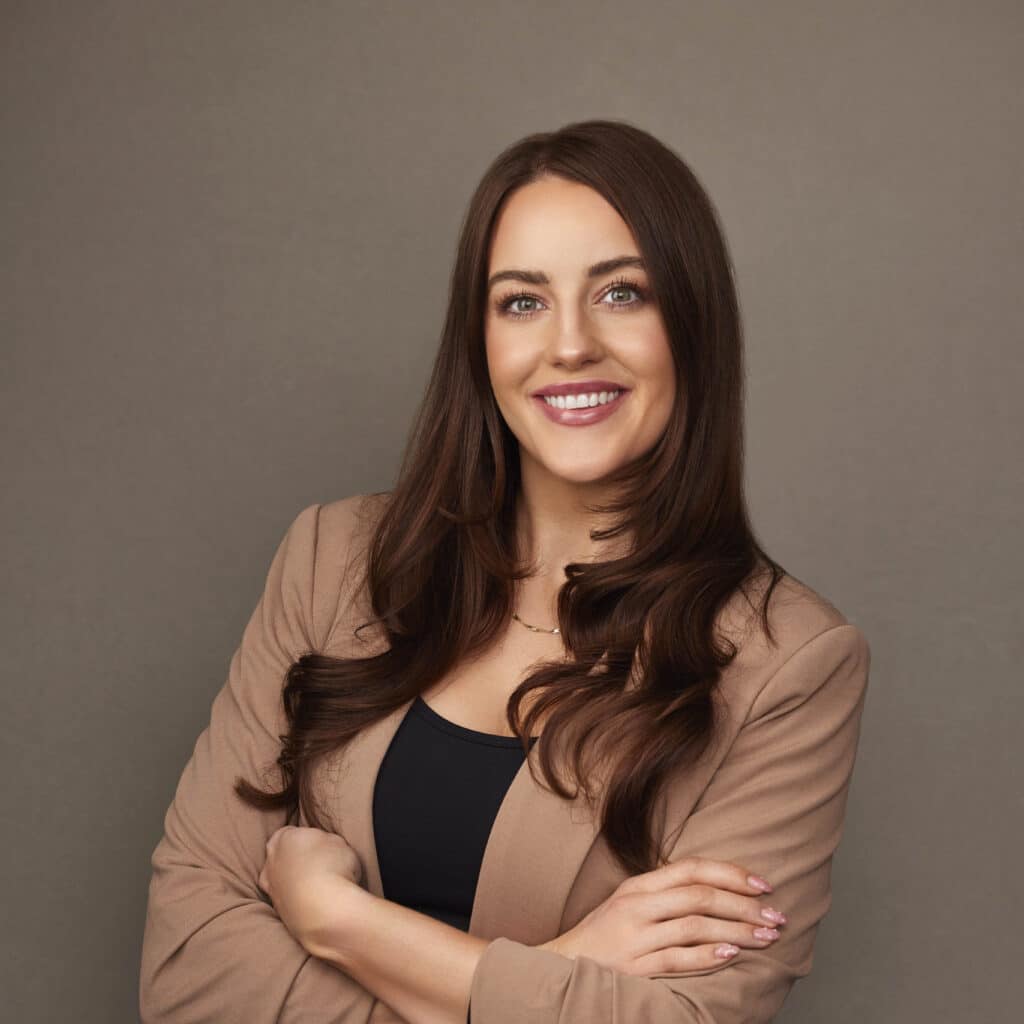 A professional portrait of a smiling woman with long, brown hair, wearing a beige blazer over a black top, arms crossed, against a gray background.