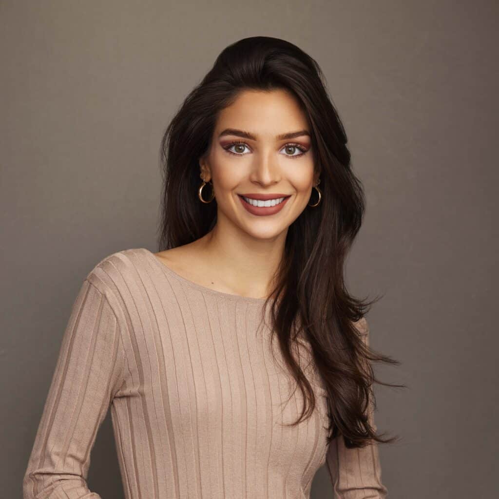 A smiling woman with long dark hair wearing a beige top and hoop earrings against a gray background. she looks directly at the camera with a friendly expression.
