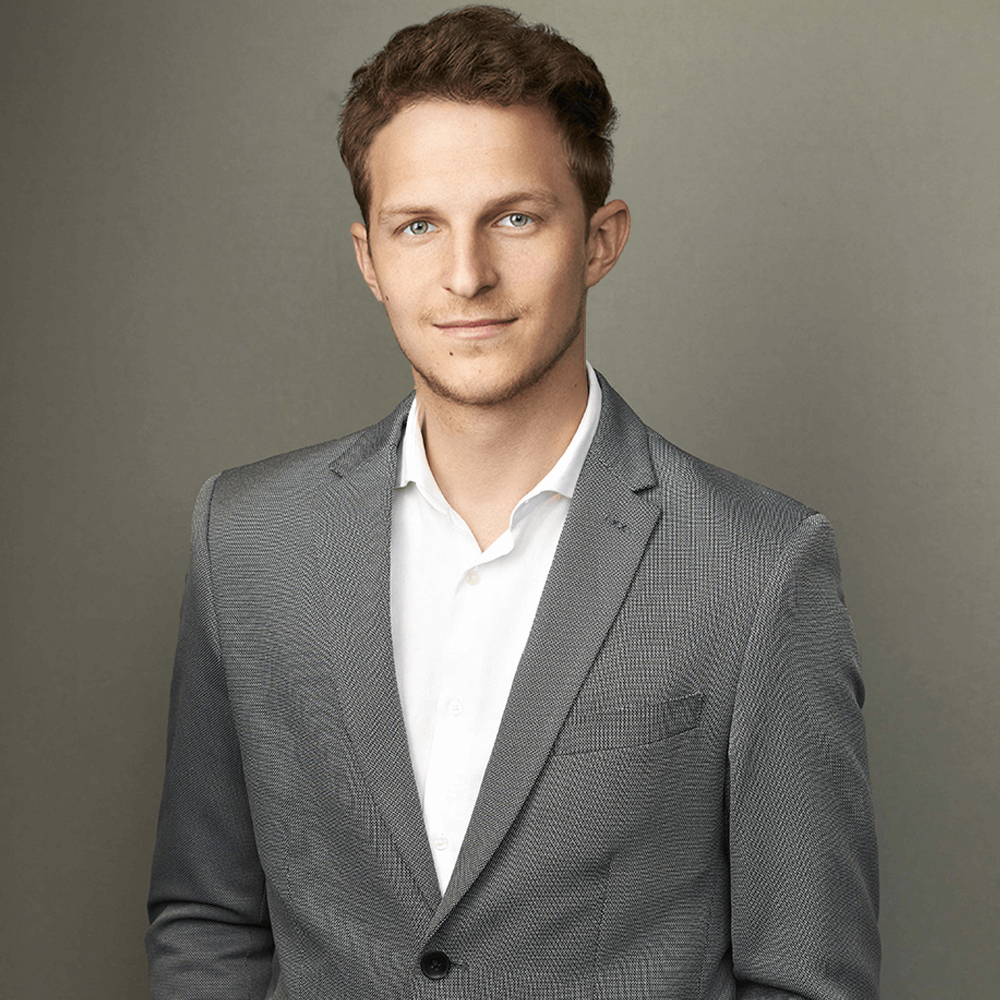 A professional portrait of a smiling young man dressed in a gray suit and white shirt, against a muted gray background. he has short, curly brown hair and a confident, friendly expression.