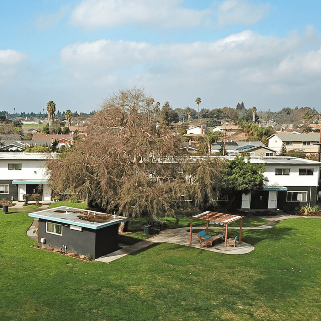 Aerial view of a suburban neighborhood featuring a large tree and a house with a flat roof and an adjacent gazebo, surrounded by green lawns and other residential buildings under a partly cloudy sky.