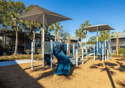 A children's playground featuring a blue slide and climbing structures, shaded by large fabric canopies, set against a backdrop of trees and residential buildings.