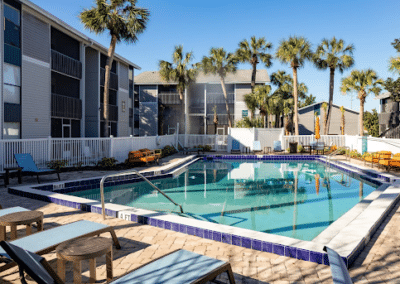 A sunny outdoor swimming pool at a residential complex with surrounding apartment buildings, deck chairs, and lush palm trees under a clear blue sky.