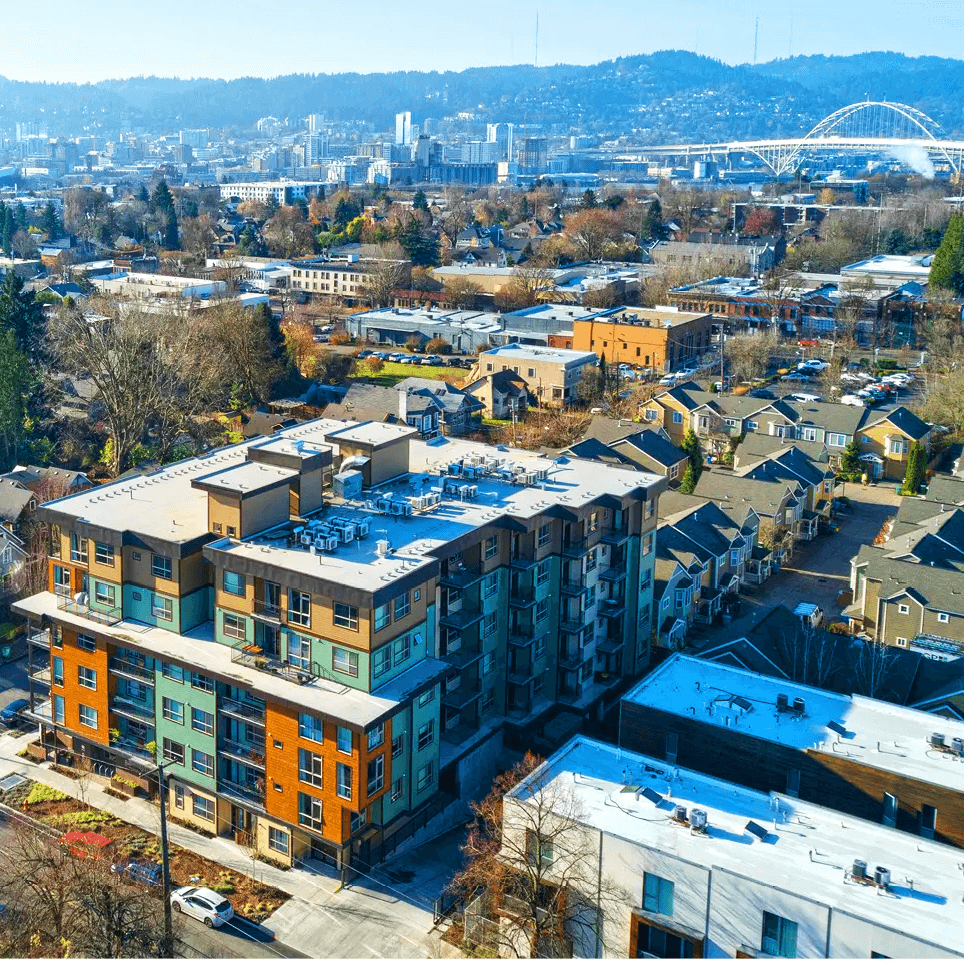 Aerial view of a modern residential apartment complex with colorful facades in a suburban setting, surrounded by other buildings and trees, with a bridge in the background under a clear blue sky.