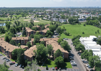 Aerial view of a residential area with multi-story brown buildings, surrounded by green spaces, trees, and a distant pond on a sunny day.