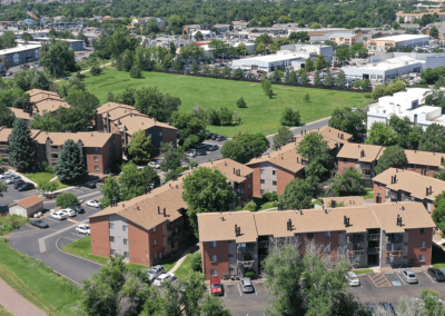 Aerial view of a suburban area featuring several brown multi-story apartment buildings surrounded by lush greenery, roads, and nearby commercial buildings.