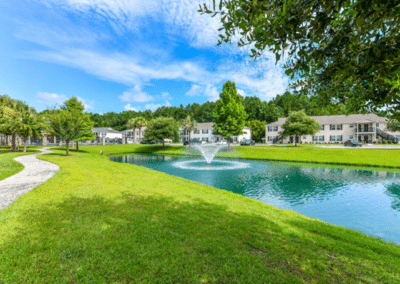 A tranquil suburban landscape featuring a pathway beside a pond with a fountain, surrounded by green grass and white townhouses under a clear blue sky.