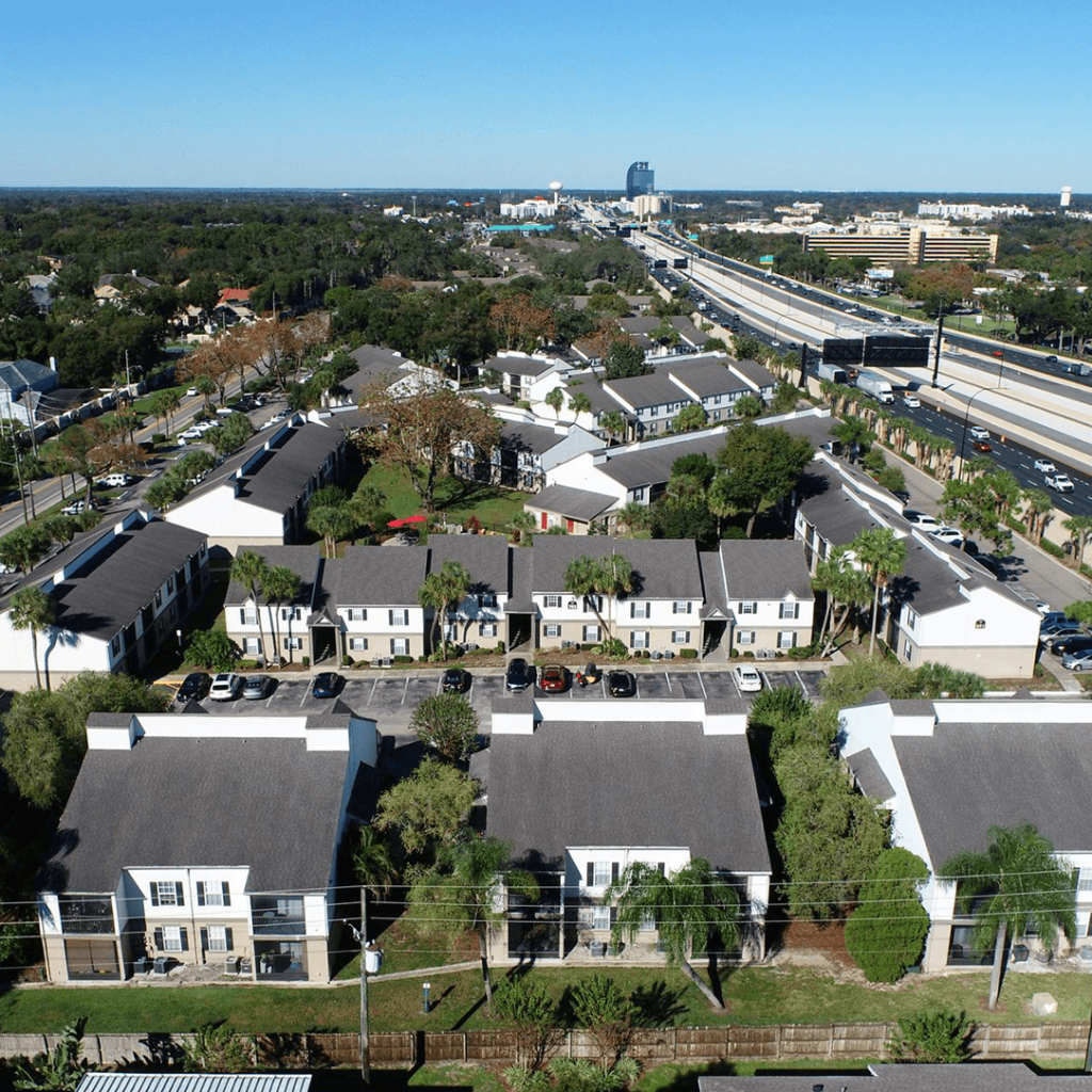 Aerial view of a residential area with uniform two-story houses alongside a busy multi-lane highway, with clear skies and scattered greenery.