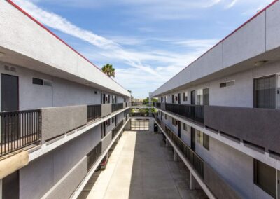 Symmetrical view of an outdoor corridor in a modern apartment complex, showing parallel walkways on upper floors framed by white walls with windows, under a clear blue sky.