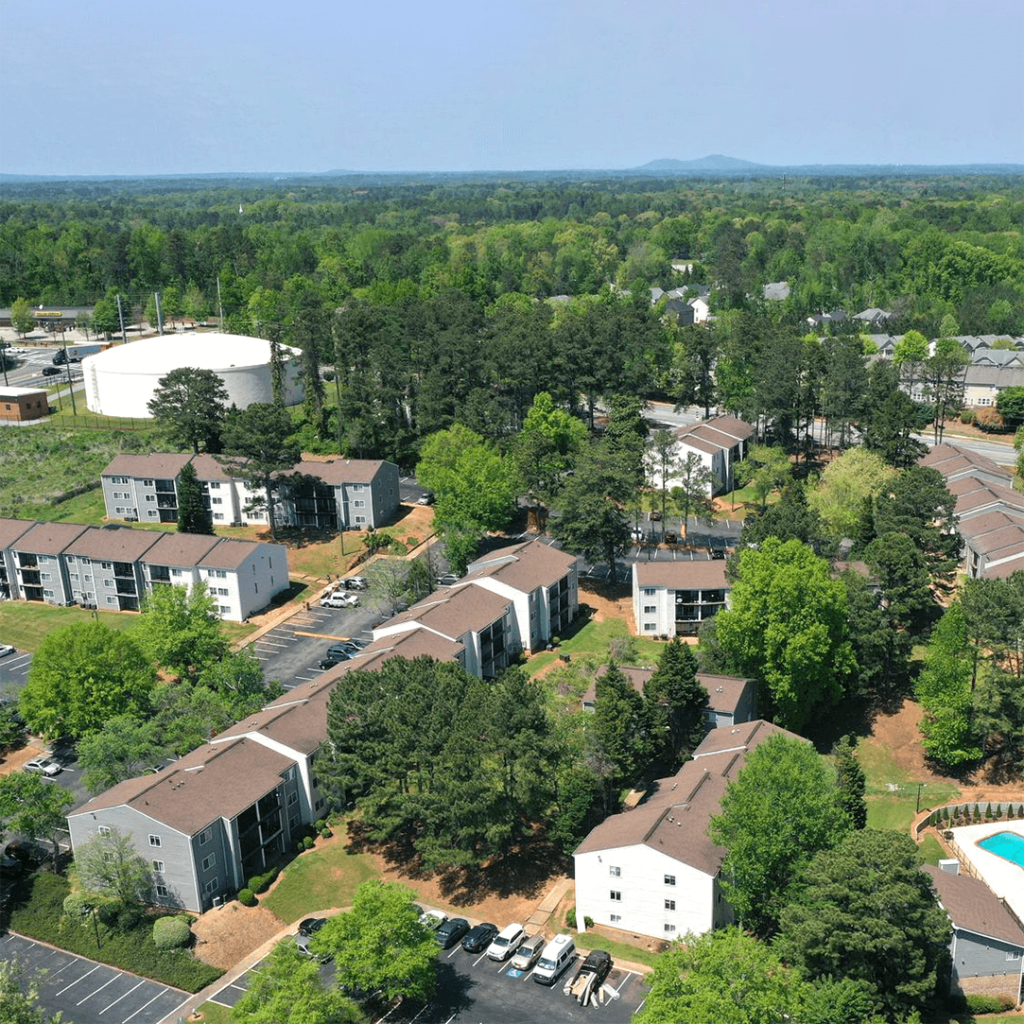 Aerial view of a suburban apartment complex surrounded by trees, featuring several two-story buildings, a swimming pool, and parked cars, under a clear sky.