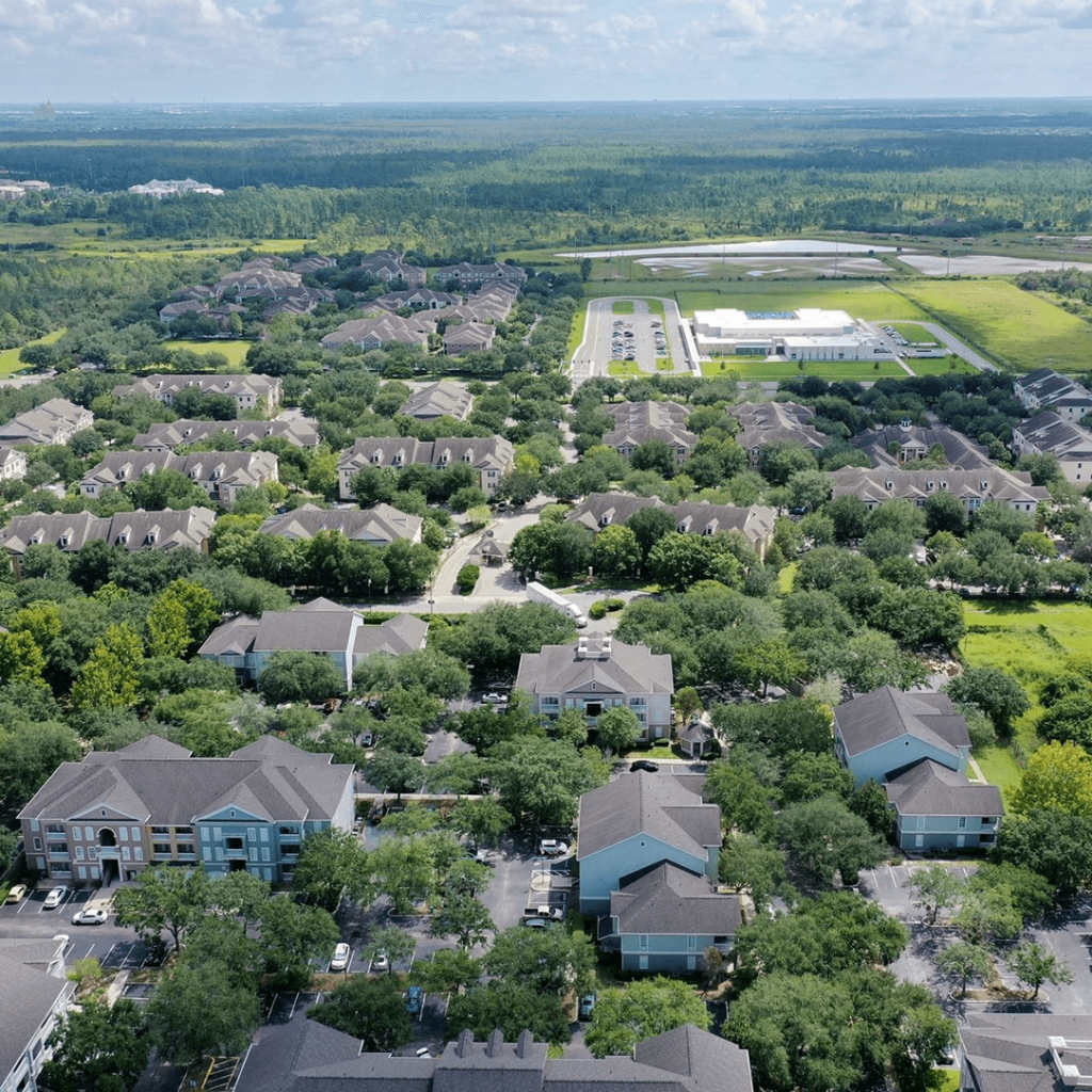 Aerial view of a residential area with a mixture of single-family homes and multi-story apartment buildings, surrounded by dense greenery and a large commercial building in the distance.