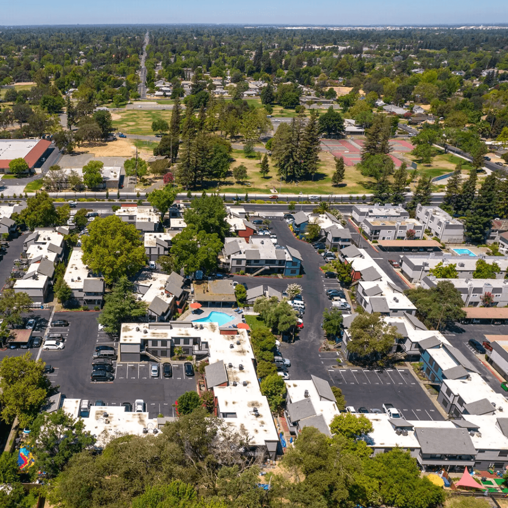 Aerial view of a suburban area featuring rows of white buildings with dark roofs, parking lots, and patches of greenery, with a large park visible in the background.