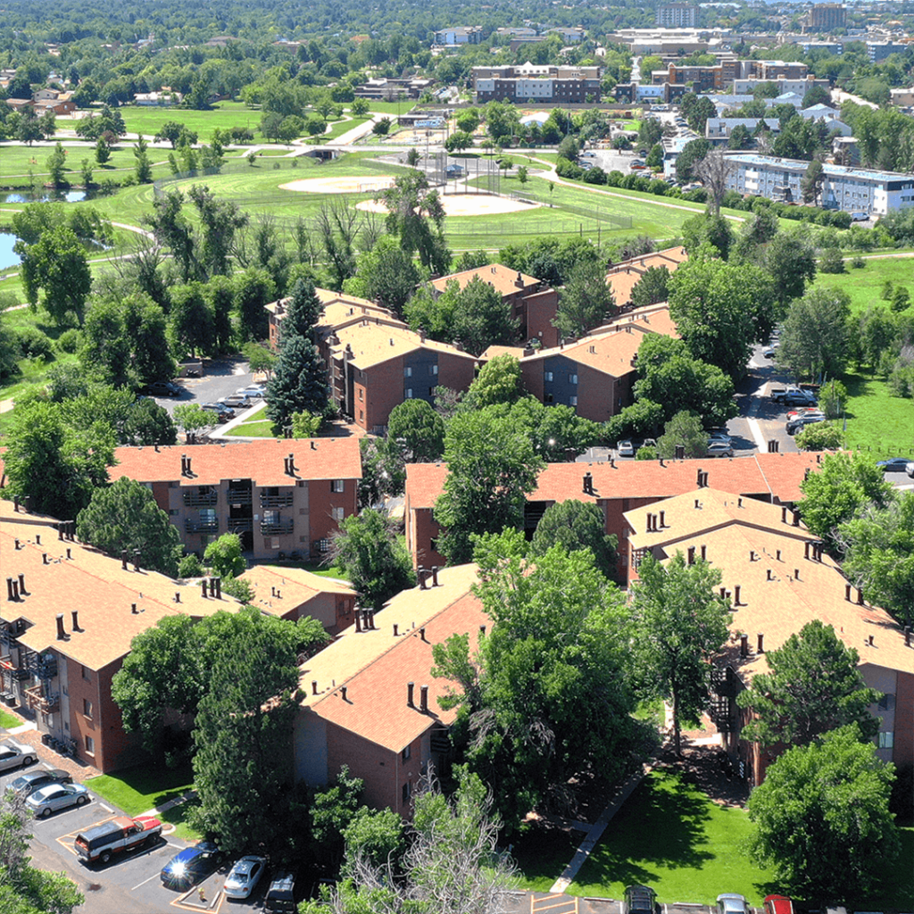 Aerial view of a residential complex with terracotta rooftops surrounded by lush green trees and a park nearby, under a clear blue sky.