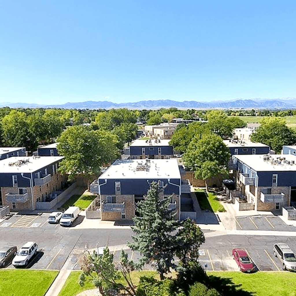 Aerial view of a suburban apartment complex with blue roofs, surrounded by greenery and parked cars, with mountains in the distance.