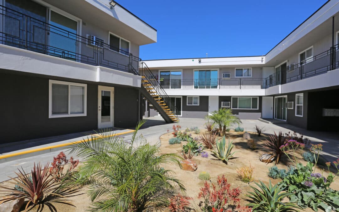 Bright, modern apartment complex with two levels featuring white and gray walls, yellow stair rails, and a landscaped courtyard with a variety of colorful plants and shrubs.