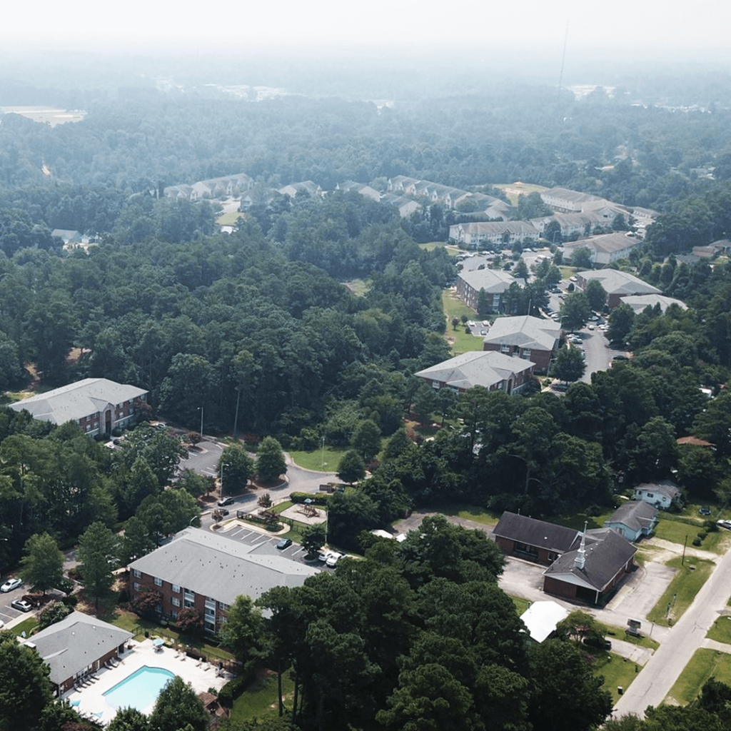 Aerial view of a suburban area with apartment buildings surrounded by trees, featuring a swimming pool and parking lots, under a hazy sky.